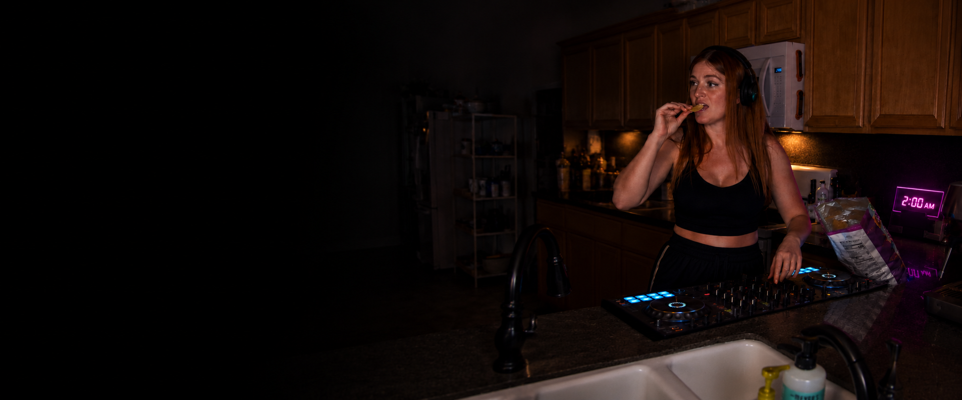 Female DJ eating potato chips in a kitchen at 2 AM while mixing music on a DJ controller, illustrating late-night snacking habits, irregular schedules, and nutrition challenges for DJs, musicians, and nightlife professionals.